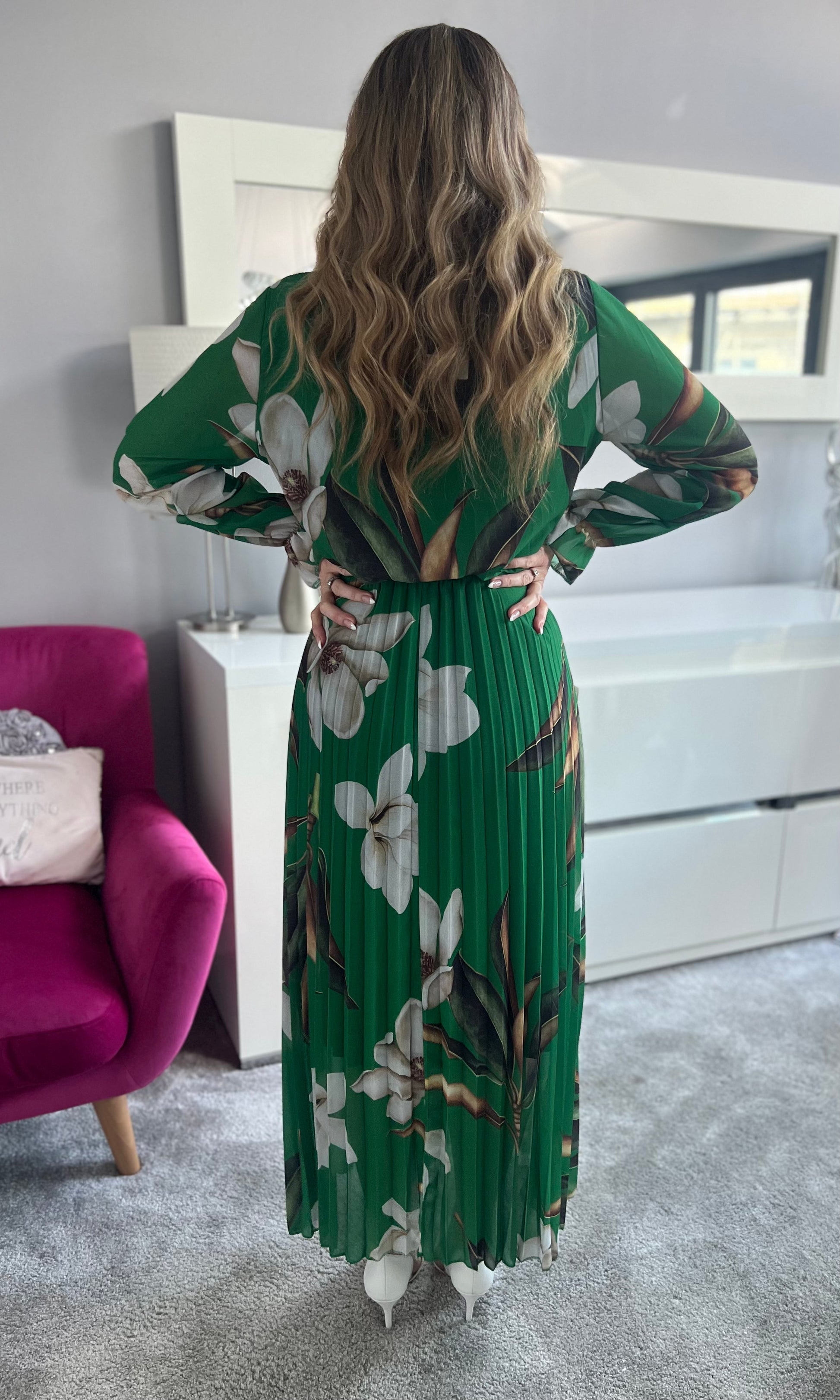 Frau in langem, grünem Kleid mit weißen Blumen, steht im modernen Wohnzimmer. Elegantes, florales Design, welliges Haar, stilvolle Einrichtung.