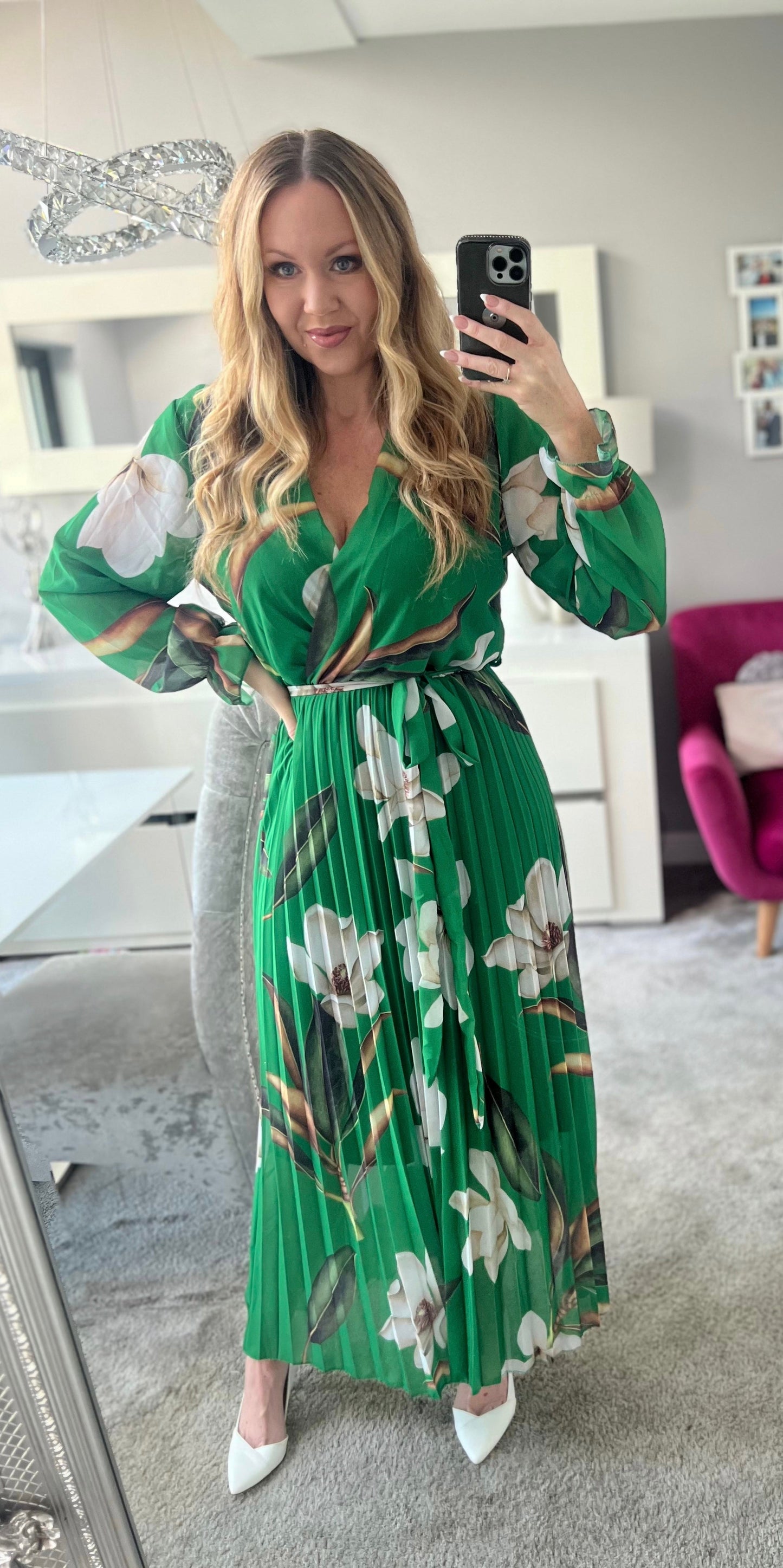 Frau in grünem, geblümtem Kleid macht Selfie im modernen Wohnzimmer. Elegantes Outfit, lange blonde Haare, stilvolle Inneneinrichtung.