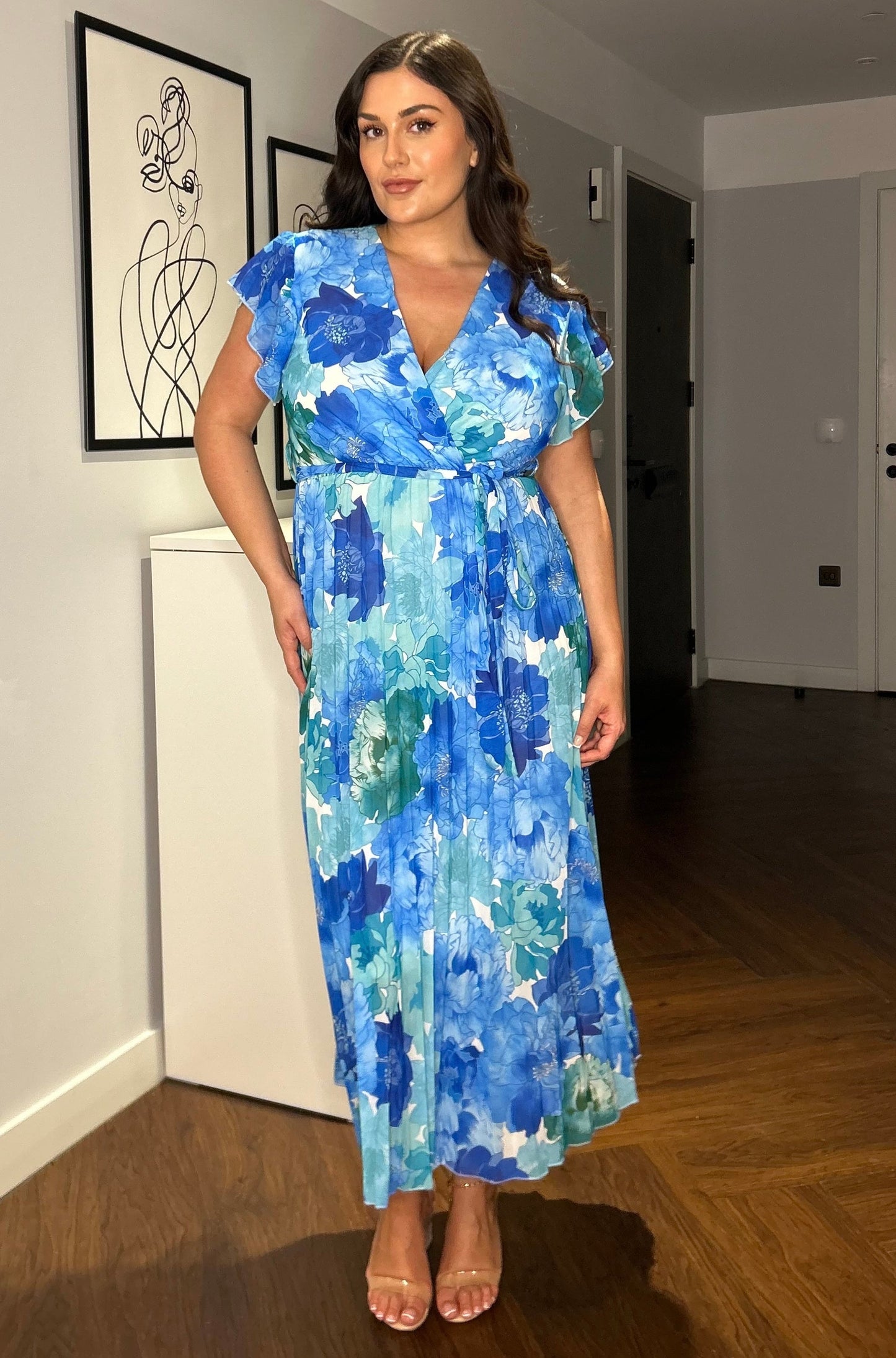 Frau in blauem, geblümtem Kleid steht in modernem Raum. Elegantes Sommerkleid, Plus-Size-Mode, florales Design, stilvolle Damenbekleidung.