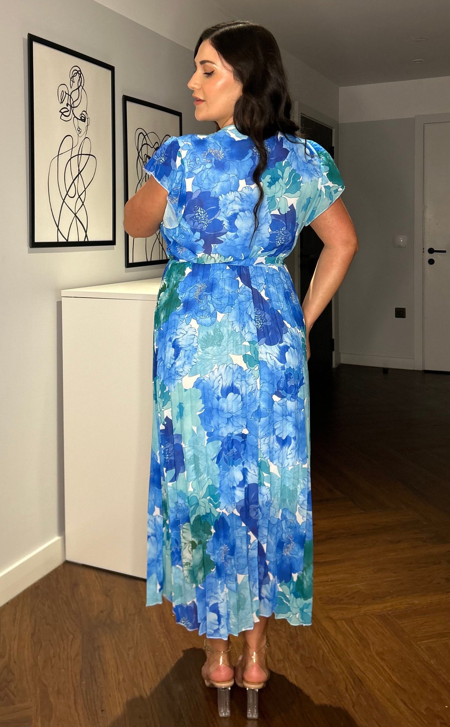 Frau in blauem, geblümtem Kleid steht in modernem Raum mit Holzboden und abstrakter Kunst an der Wand. Elegantes Sommeroutfit, Mode, Inneneinrichtung.