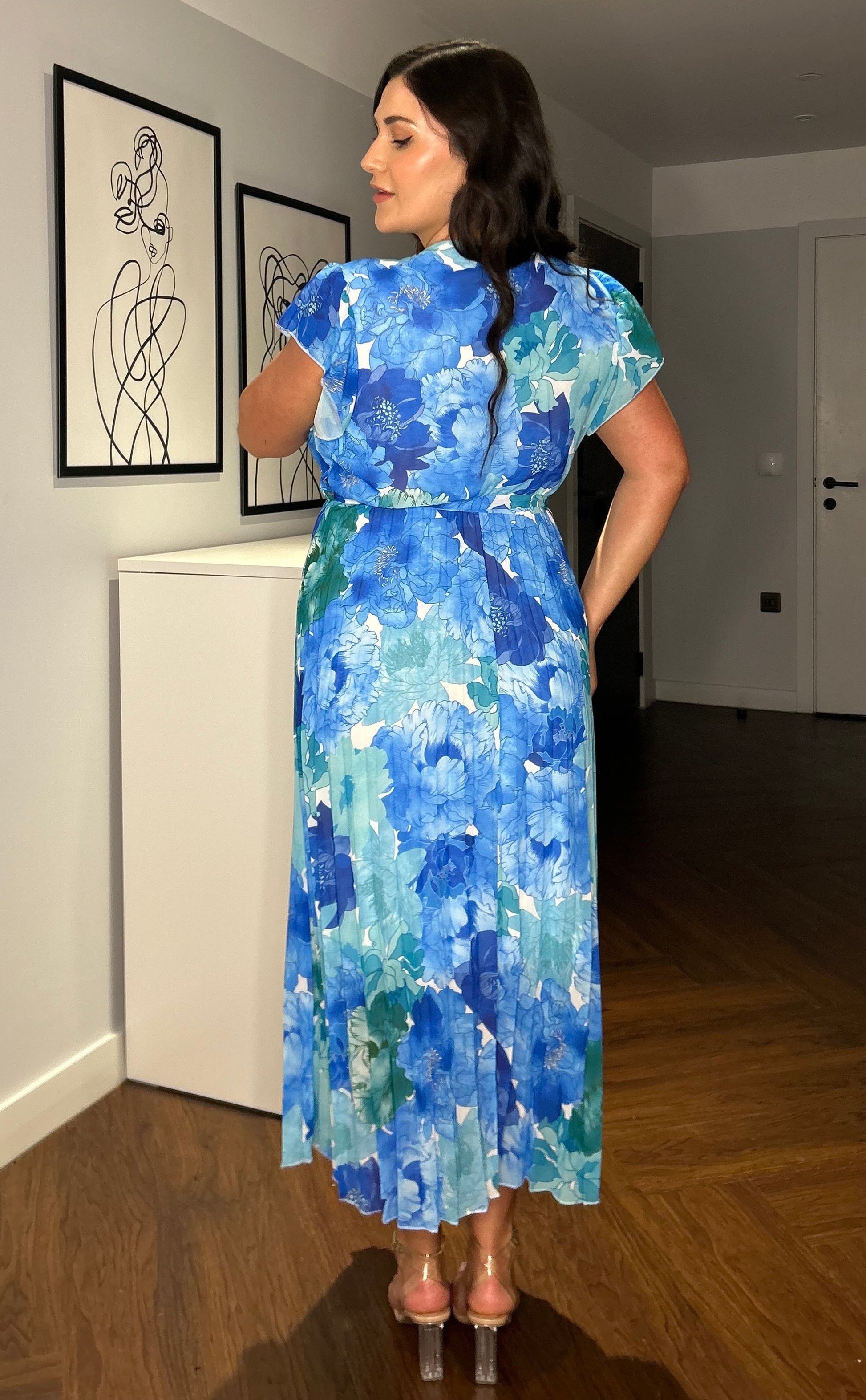 Frau in blauem, geblümtem Kleid steht in modernem Raum mit Holzboden und abstrakter Kunst an der Wand. Elegantes Sommeroutfit, Mode, Inneneinrichtung.