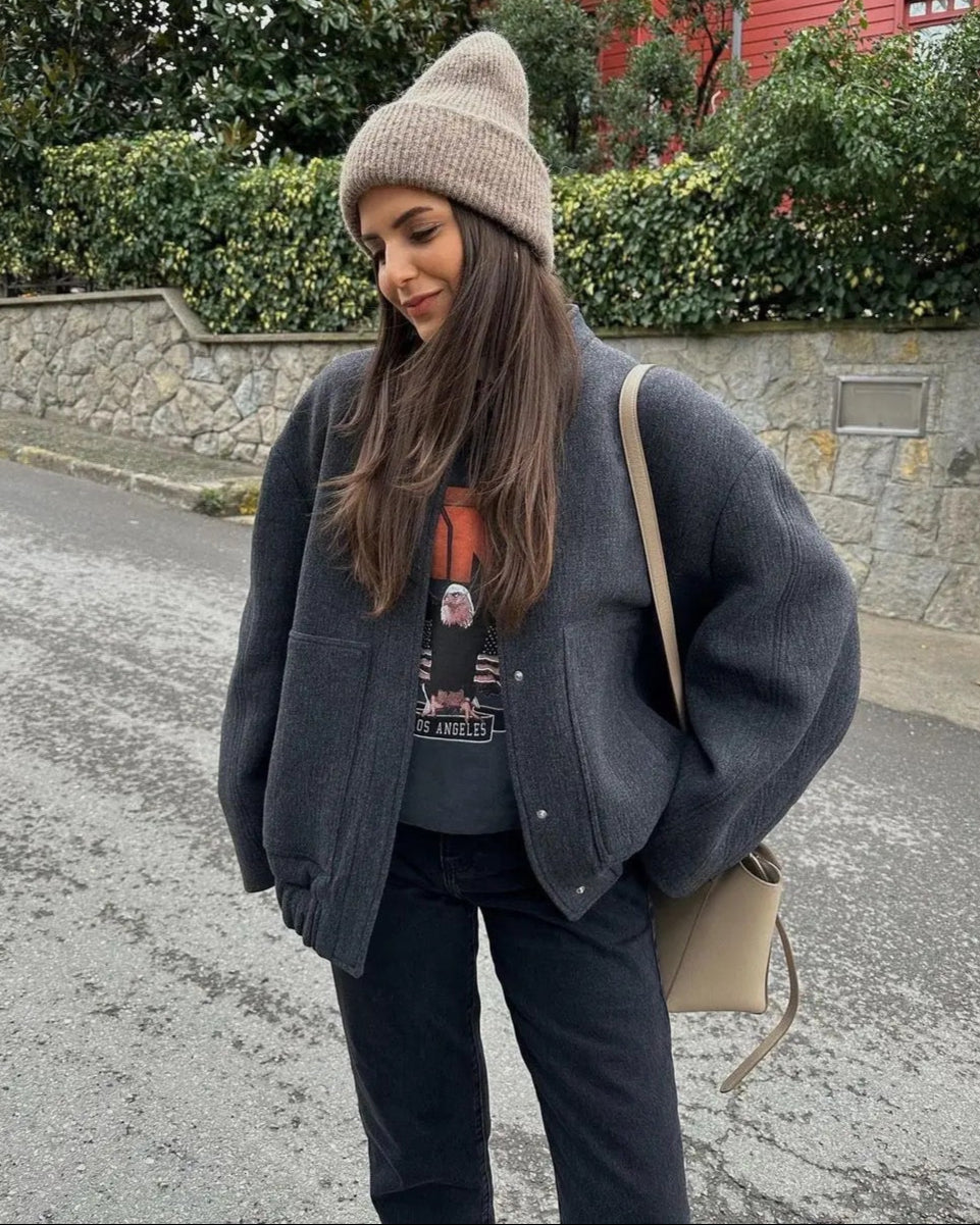 Frau in grauer Jacke und Mütze auf Straße, trägt schwarze Jeans und T-Shirt, mit beiger Tasche. Wintermode, lässiger Stil, Outdoor-Kleidung.