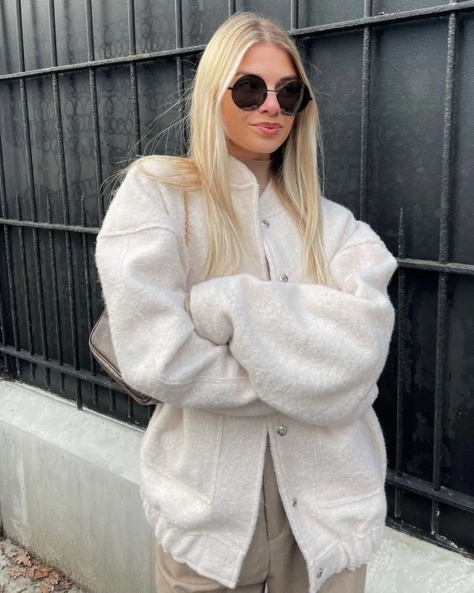Blonde Frau mit Sonnenbrille trägt weiße, flauschige Jacke vor schwarzem Gitter. Modetrend, Herbstmode, Streetstyle, lässiger Look.