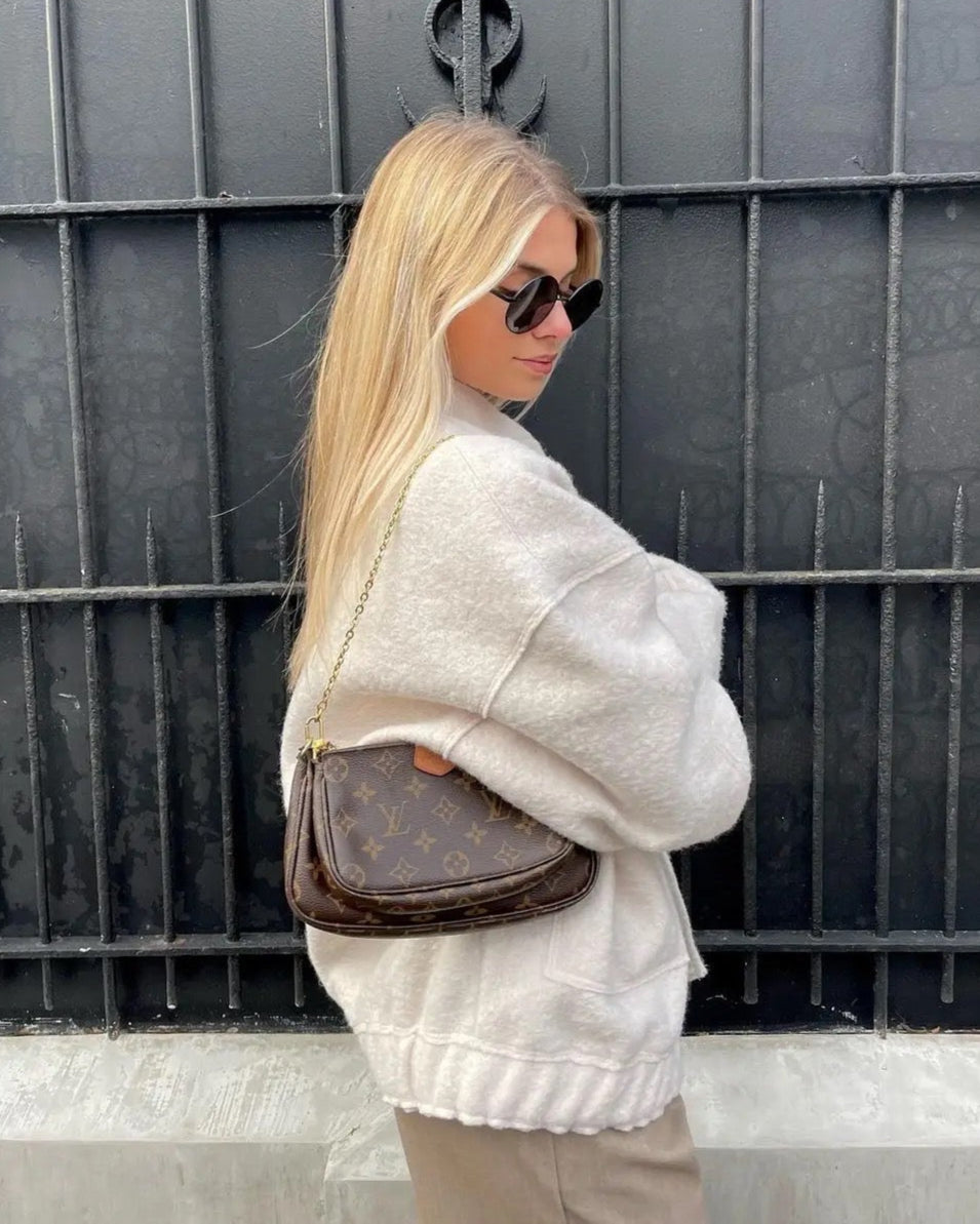 Blonde Frau mit Sonnenbrille trägt eine braune Louis Vuitton Handtasche vor einem schwarzen Gitter. Elegante Mode, Luxusaccessoire, Streetstyle.