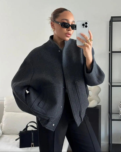 Frau in übergroßer grauer Jacke, schwarze Sonnenbrille, hält iPhone, steht vor weißer Wand. Moderne Mode, Selfie, stilvoller Look.