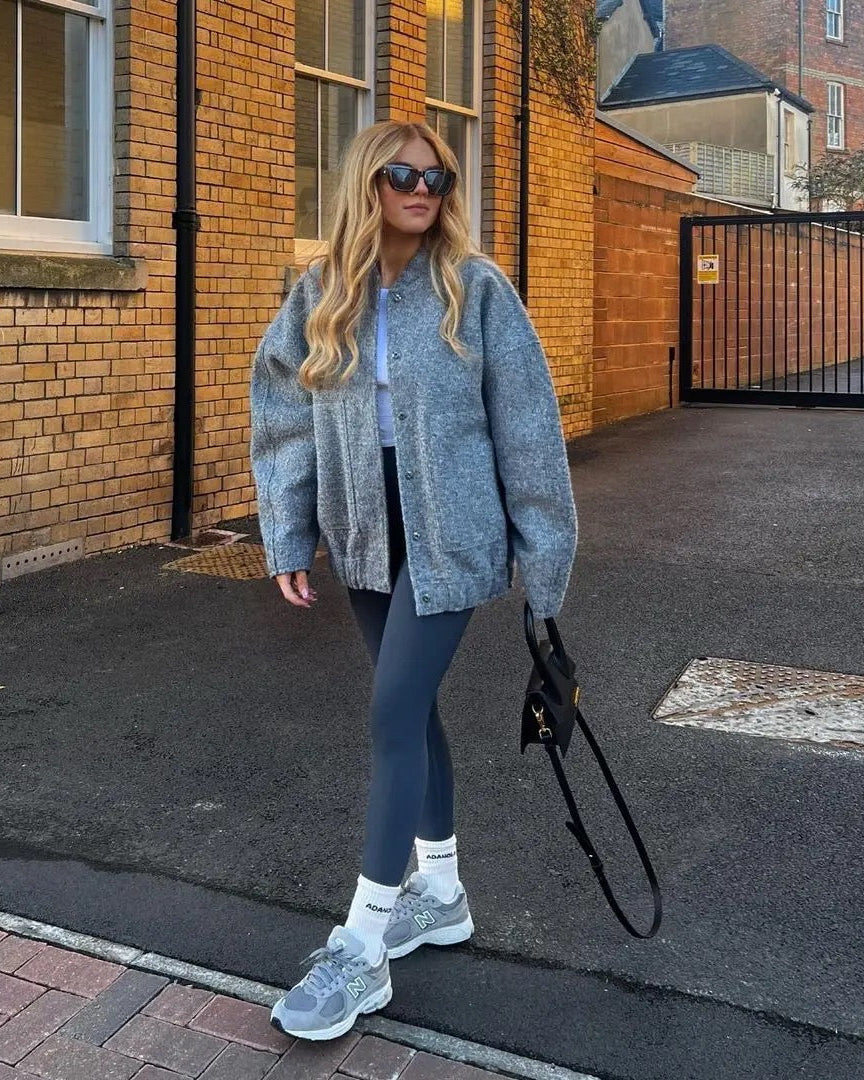 Frau in grauer Strickjacke und Leggings, trägt Sonnenbrille und Sneakers, hält schwarze Tasche. Urbaner Hintergrund mit Backsteinmauer.