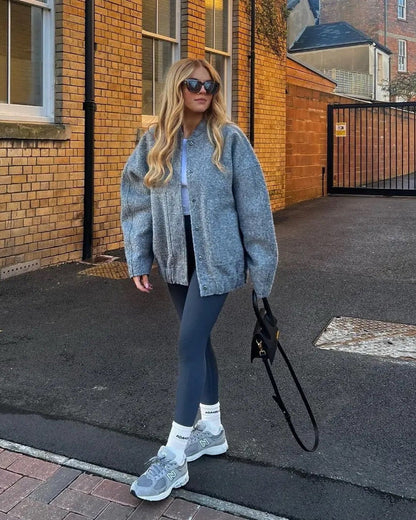 Frau in grauer Strickjacke und Leggings, trägt Sonnenbrille und Sneakers, hält schwarze Tasche. Urbaner Hintergrund mit Backsteinmauer.