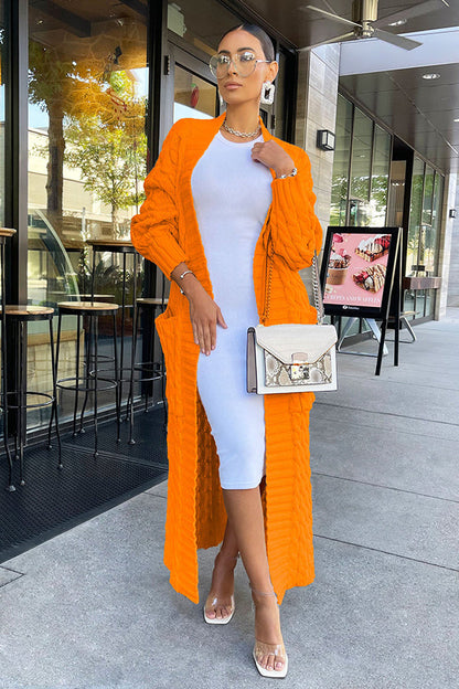 Frau in weißem Kleid und orangefarbenem Cardigan, trägt Sonnenbrille und Handtasche, steht vor Café. Elegante Mode, Streetstyle, Sommeroutfit.
