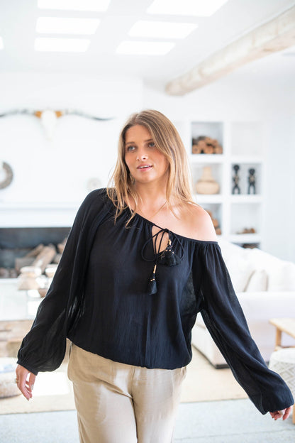 Junge Frau in schwarzer Bluse und beigen Hosen steht in modernem Wohnzimmer mit heller Einrichtung und dekorativen Elementen im Hintergrund.