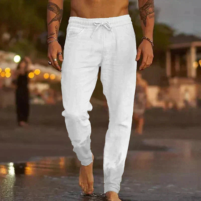 Ein Mann in weißen Leinenhosen spaziert barfuß am Strand entlang. Sommermode, Freizeitkleidung, Strandspaziergang, Herrenmode.
