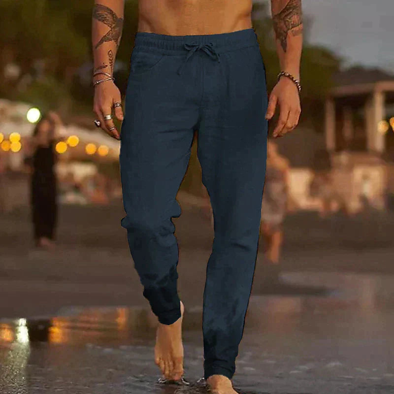Mann in blauer Jogginghose geht barfuß am Strand entlang bei Sonnenuntergang, Freizeitmode, Sommerabend, entspannte Atmosphäre.