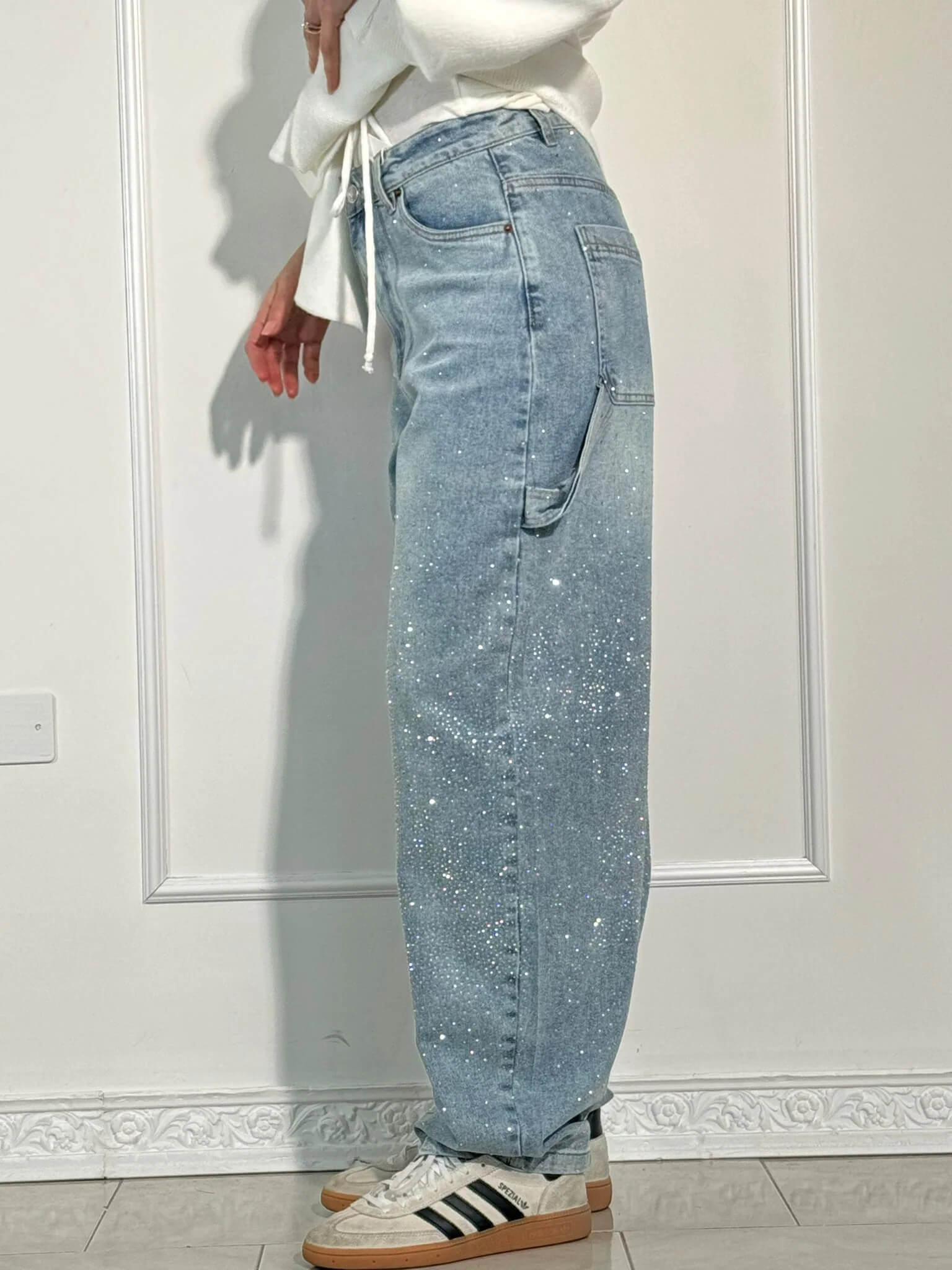 Damen-Jeans mit weitem Bein, hellblau, funkelnde Details, kombiniert mit weißen Sneakers und einem weißen Oberteil, vor weißer Wand.