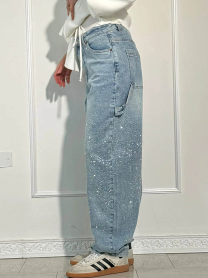 Damen-Jeans mit weitem Bein, hellblau, funkelnde Details, kombiniert mit weißen Sneakers und einem weißen Oberteil, vor weißer Wand.