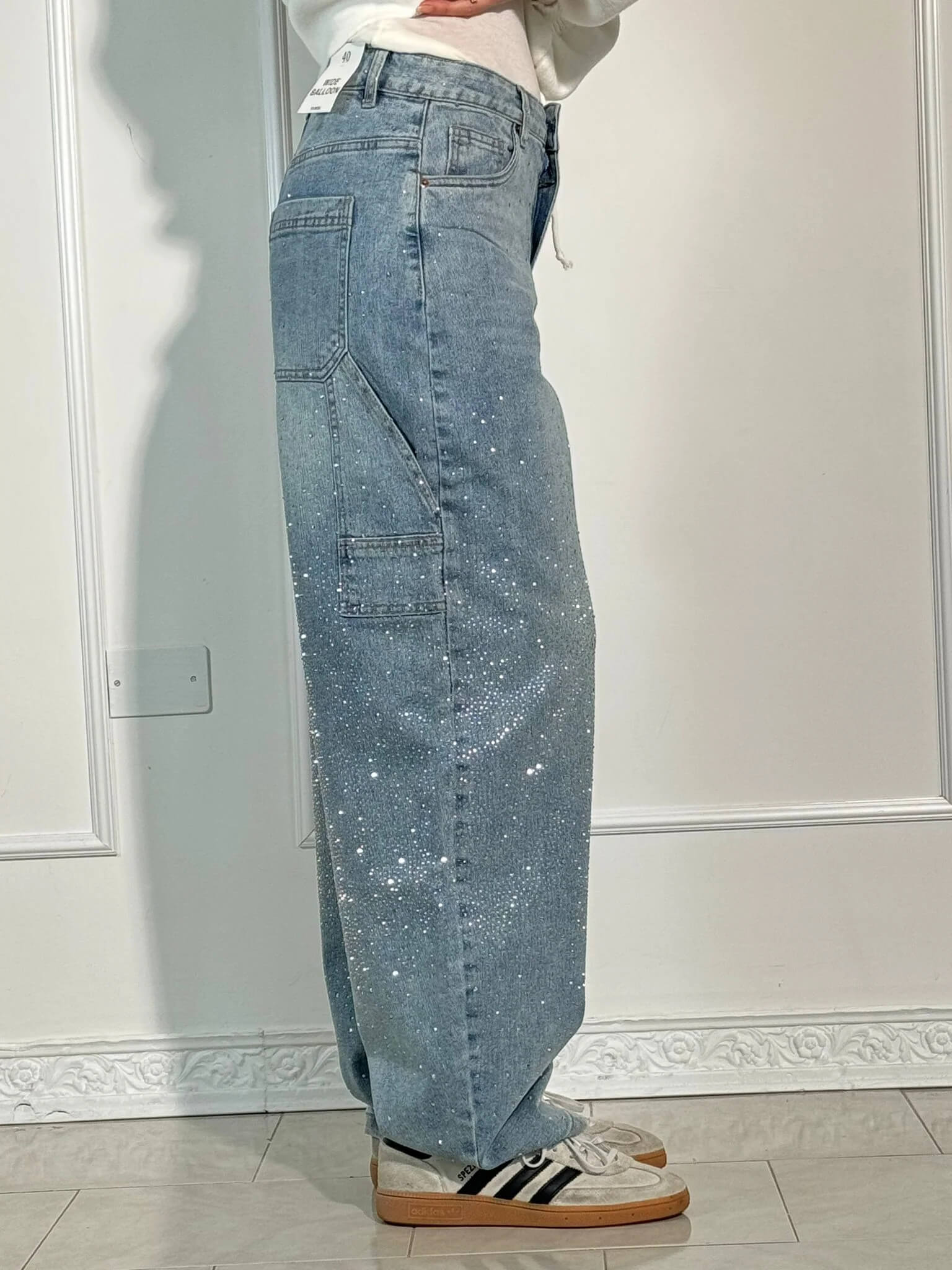 Damen-Jeans mit weitem Bein, hellblau, glitzernd, kombiniert mit weißen Sneakers und schwarzen Streifen, vor weißer Wand.