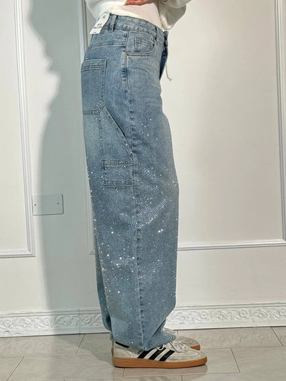 Damen-Jeans mit weitem Bein, hellblau, glitzernd, kombiniert mit weißen Sneakers und schwarzen Streifen, vor weißer Wand.
