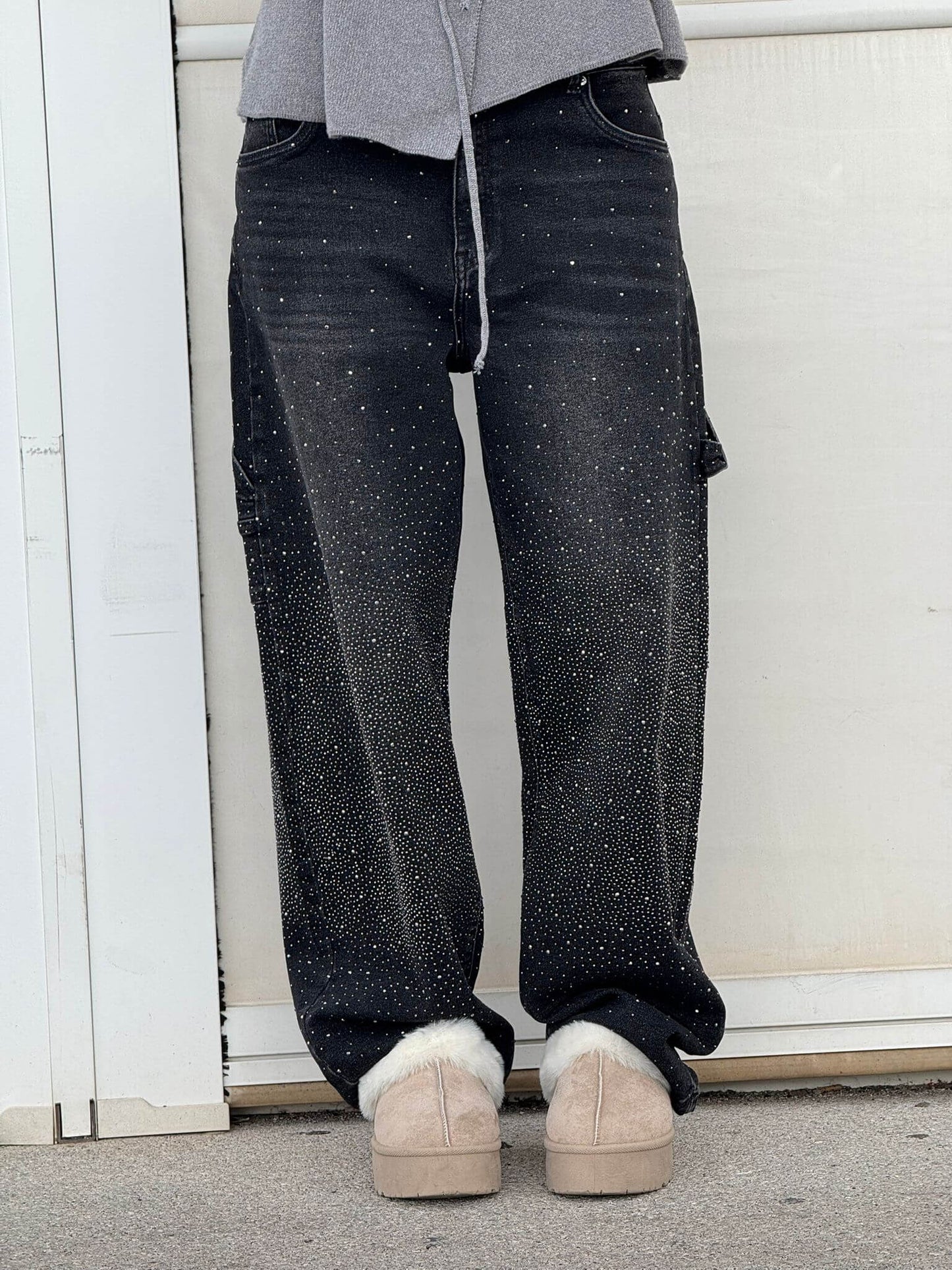 Graue Jogginghose, schwarze Jeans mit Glitzerpunkten, beige Hausschuhe. Lässiger Streetwear-Look, modisch und bequem.