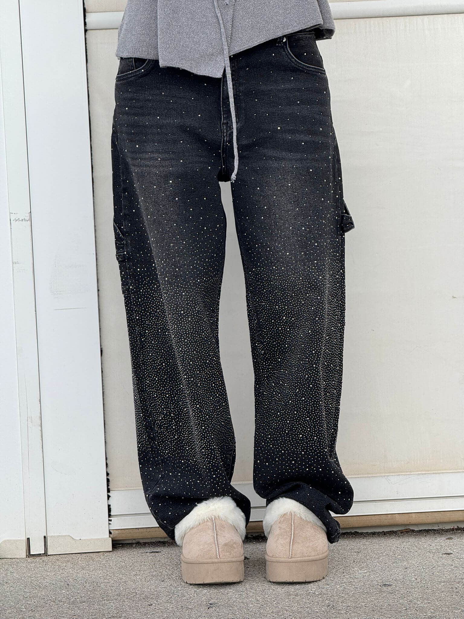 Graue Jogginghose, schwarze Jeans mit Glitzerpunkten, beige Hausschuhe. Lässiger Streetwear-Look, modisch und bequem.