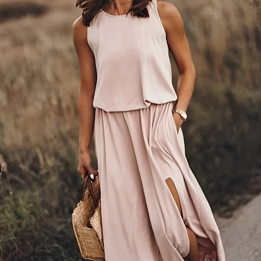 Frau in einem eleganten, ärmellosen, beigen Maxikleid mit Schlitz, hält eine Strohtasche, steht auf einem ländlichen Weg. Sommermode, Damenkleidung.