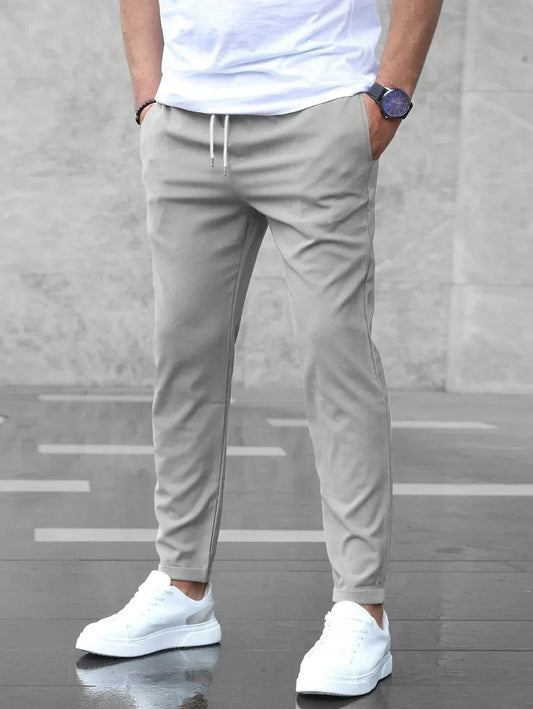 Mann in grauer Jogginghose und weißen Sneakers, trägt weißes T-Shirt, steht auf nassem Boden. Moderne Herrenmode, lässiger Stil.