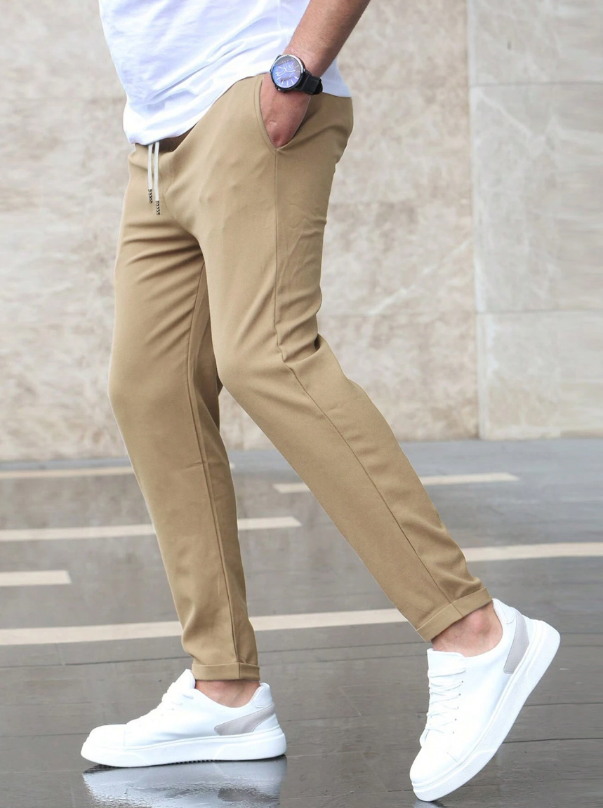 Mann in beigen Chino-Hosen und weißen Sneakers, seitlich gehend auf grauem Boden. Lässiger Stil, moderne Herrenmode, Outdoor-Aufnahme.
