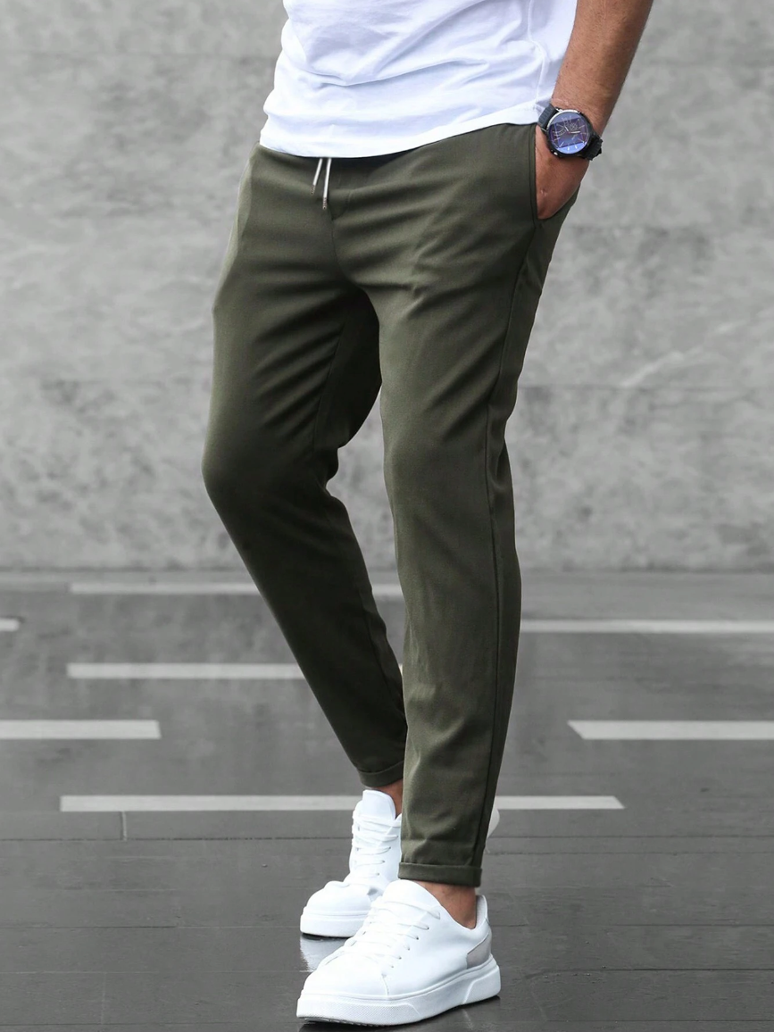 Mann in olivgrüner Jogginghose und weißen Sneakers vor grauem Hintergrund. Lässige Herrenmode, sportlicher Look, moderne Freizeitkleidung.
