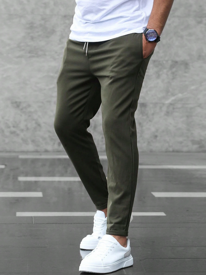 Mann in olivgrüner Jogginghose und weißen Sneakers vor grauem Hintergrund. Lässige Herrenmode, sportlicher Look, moderne Freizeitkleidung.