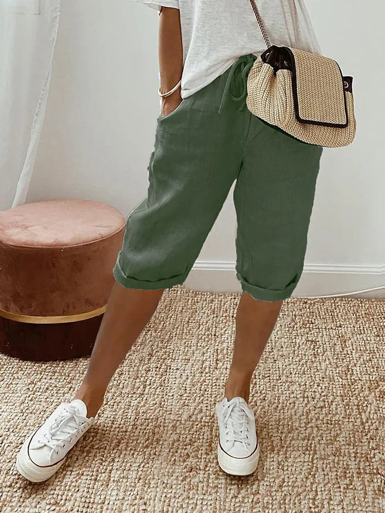 Frau in grüner Leinenhose und weißem T-Shirt, trägt eine beige Umhängetasche und weiße Sneaker, steht auf einem Teppich.