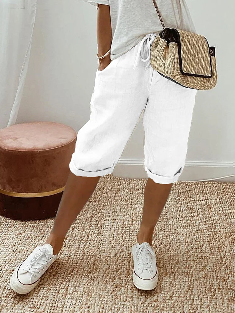 Frau in weißer Bermuda-Shorts und Sneakers, beige Umhängetasche, lässiger Look, Sommermode, Innenraum mit Teppich und Hocker.