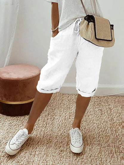 Frau in weißer Bermuda-Shorts und Sneakers, beige Umhängetasche, lässiger Look, Sommermode, Innenraum mit Teppich und Hocker.