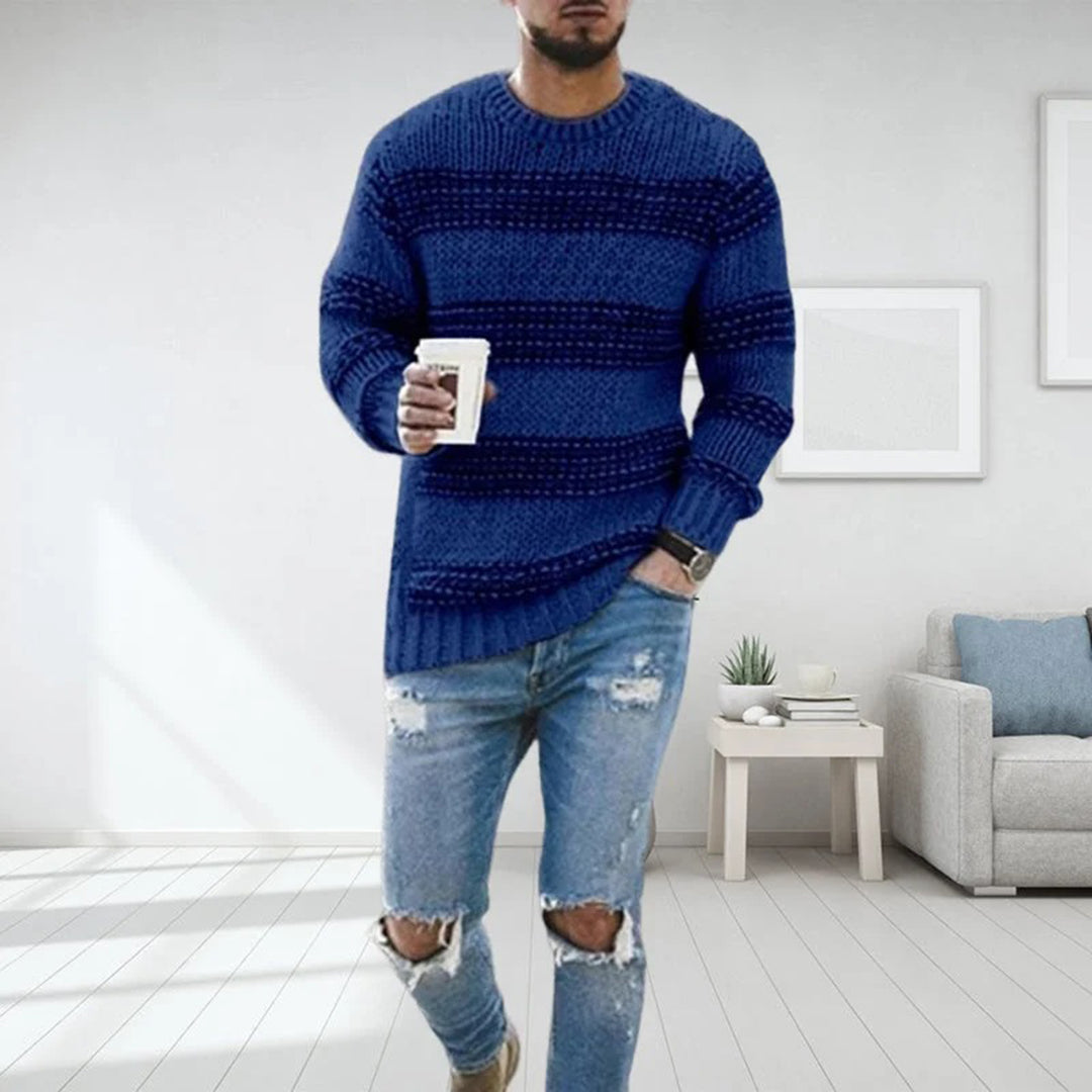Mann in blauem Strickpullover und zerrissenen Jeans hält Kaffeetasse, steht in modernem Wohnzimmer. Mode, Freizeitkleidung, Herrenoutfit.