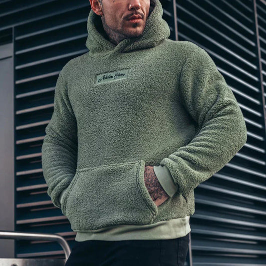 Mann in grünem Fleece-Hoodie vor moderner Architektur. Bequeme, warme Herrenmode, ideal für Herbst und Winter. Trendiger Streetwear-Look.