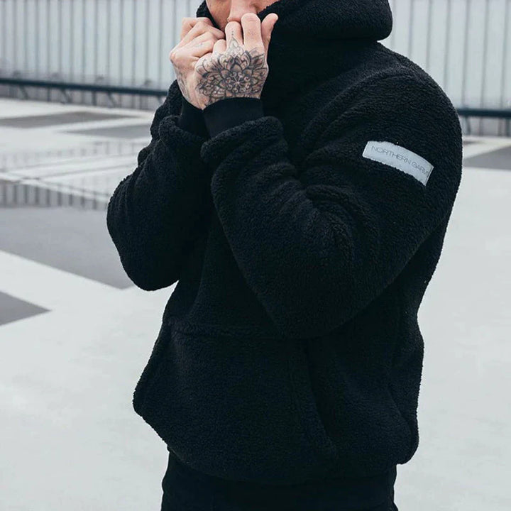 Person in schwarzem Fleece-Hoodie mit tätowierter Hand, steht auf einem urbanen Dach. Modischer Winterlook, Streetwear, Outdoor-Bekleidung.