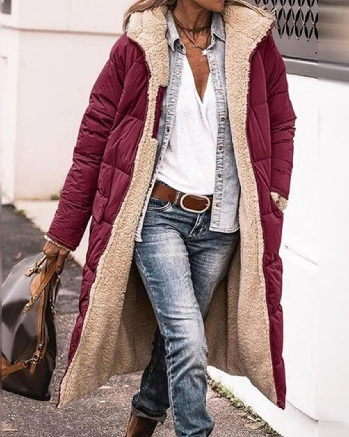 Frau in langem, weinrotem Steppmantel mit weißem Futter, kombiniert mit Jeans und weißem Shirt, spaziert auf Gehweg. Wintermode, Damenmantel, Streetstyle.