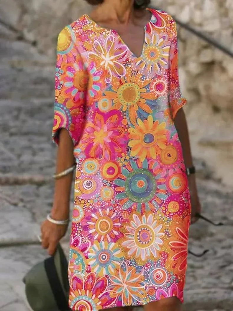 Frau in buntem, geblümtem Sommerkleid mit leuchtenden Blumenmustern, steht auf gepflastertem Weg. Modetrend, farbenfrohes Design, Freizeitkleidung.
