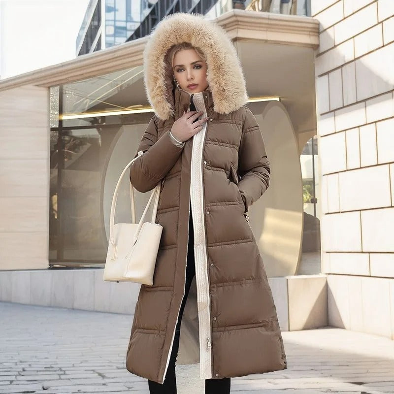 Frau in braunem Daunenmantel mit Fellkapuze, hält weiße Handtasche, steht vor moderner Architektur. Wintermode, stilvoll, elegant.