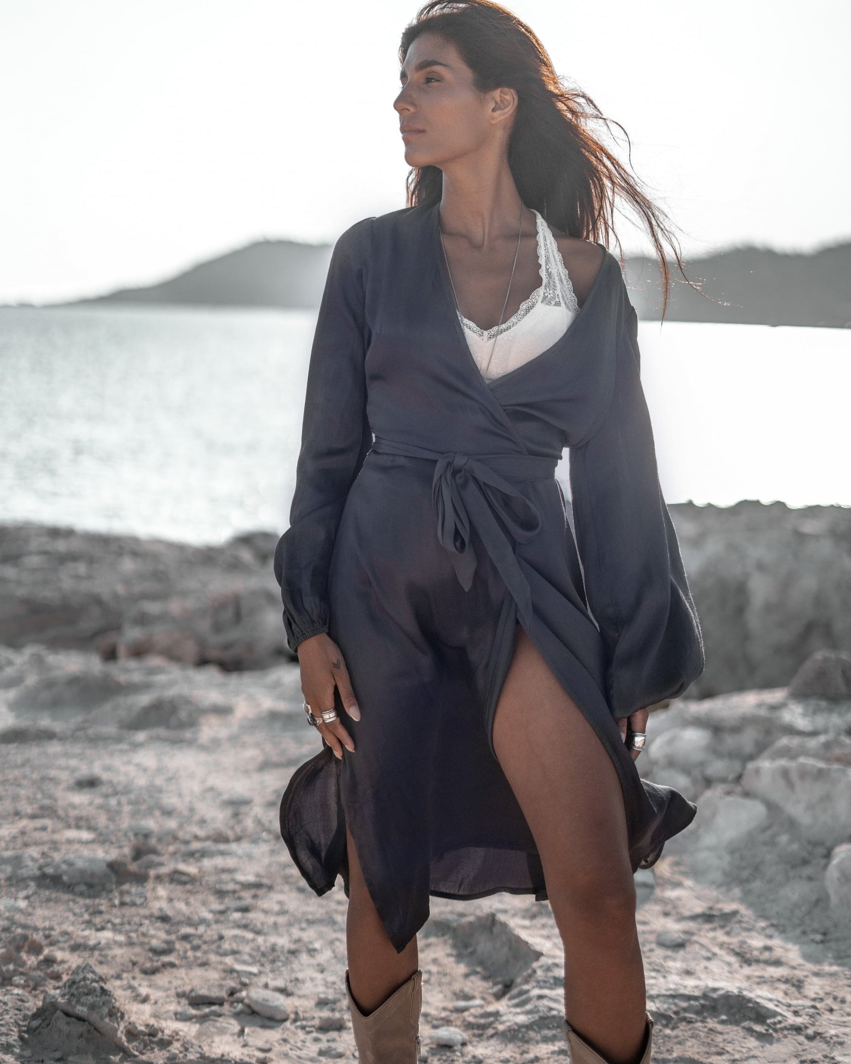 Frau in dunkelblauem Kleid und weißem Top am Strand, windiges Haar, sonniger Tag, Meer und Hügel im Hintergrund. Sommermode, Strandkleidung.