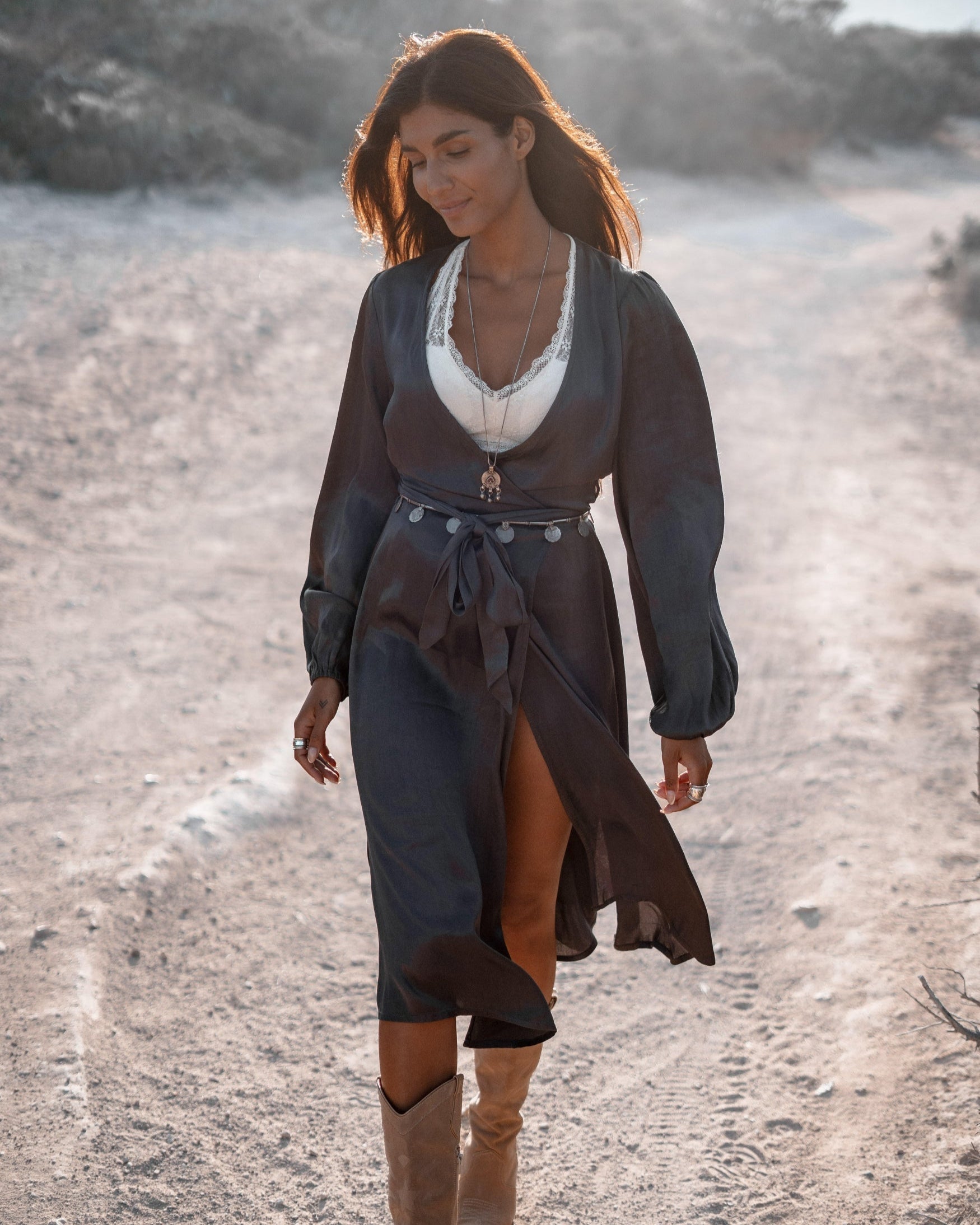 Frau in elegantem, dunklem Kleid mit Gürtel und Stiefeln auf staubigem Weg bei Sonnenuntergang. Mode, Outdoor, Boho-Stil, Herbstkleidung.
