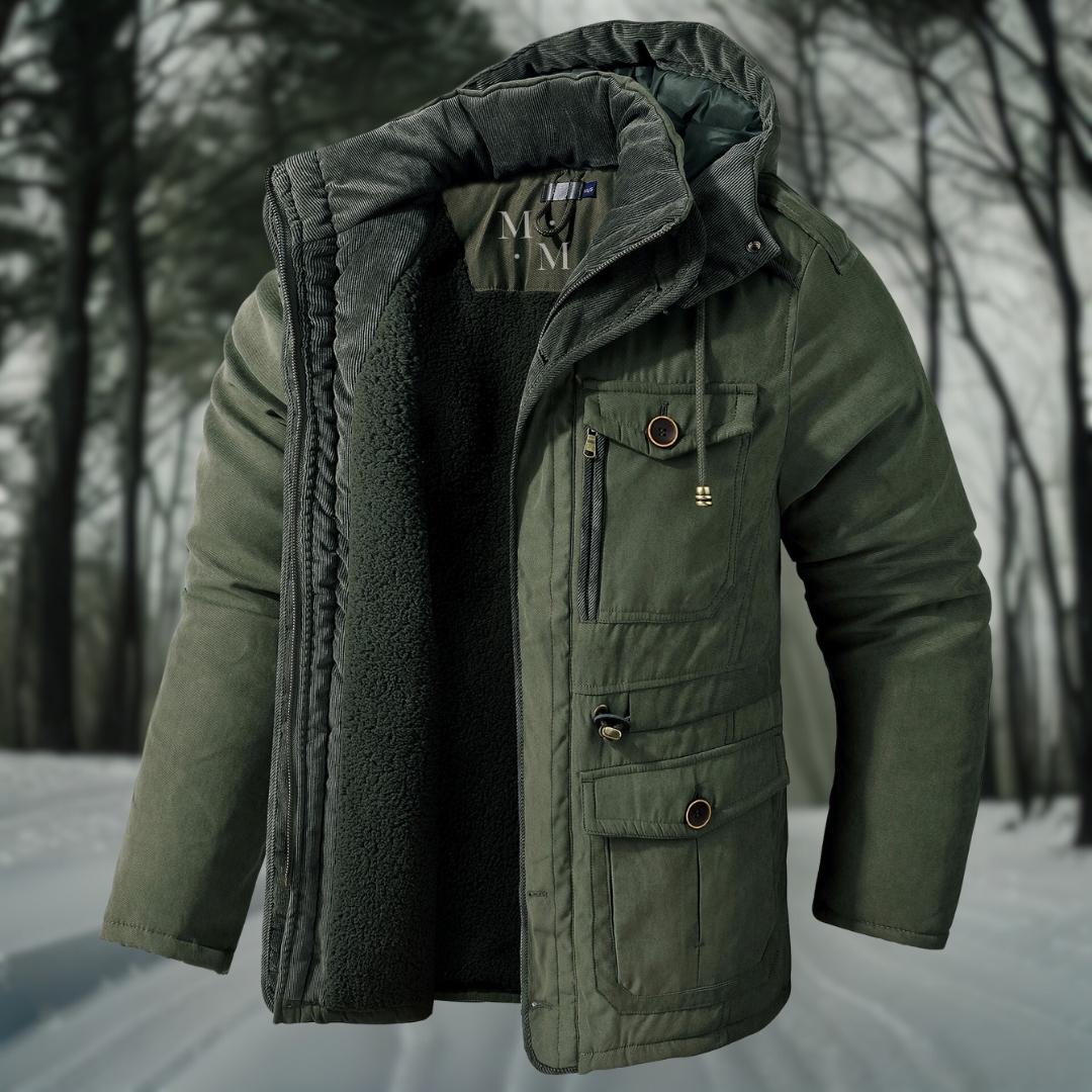 Grüne Winterjacke mit Kapuze und mehreren Taschen, gefüttert mit Fleece, vor einem verschneiten Waldhintergrund. Ideal für Outdoor-Aktivitäten.
