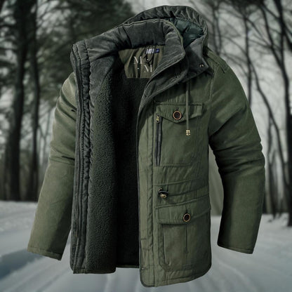Grüne Winterjacke mit Kapuze und mehreren Taschen, gefüttert mit Fleece, vor einem verschneiten Waldhintergrund. Ideal für Outdoor-Aktivitäten.