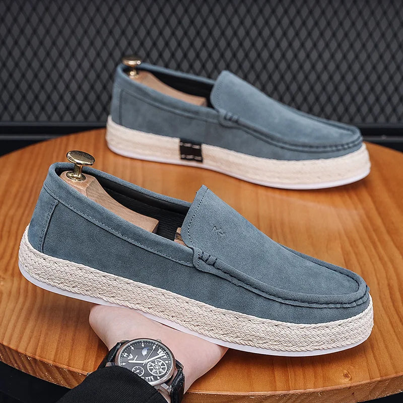 Graue Herren-Slipper aus Wildleder auf Holztisch, mit weißer Sohle und Espadrille-Details. Elegante Freizeitschuhe für stilvolle Outfits.
