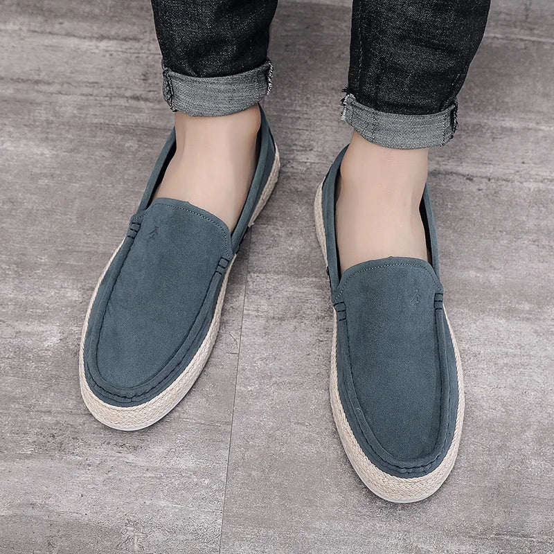 Graue Herren-Slipper aus Wildleder mit geflochtener Sohle, getragen auf grauem Boden. Bequeme, stilvolle Freizeitschuhe für den Alltag.