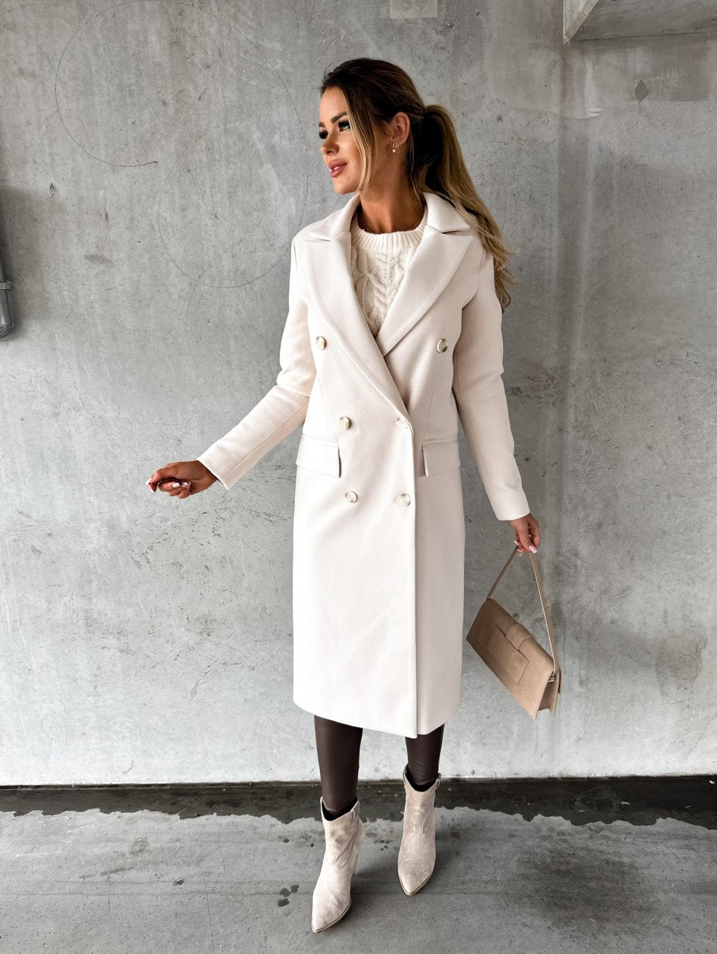 Frau in elegantem, weißem Wollmantel, kombiniert mit beiger Handtasche und Stiefeletten, vor grauer Betonwand. Wintermode, stilvoll, modern.