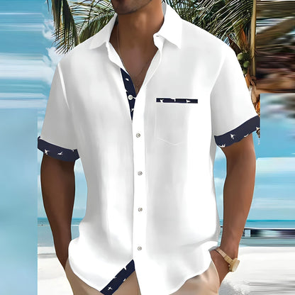 Mann in weißem Kurzarmhemd mit blauem Muster am Strand, Palmen im Hintergrund. Sommermode, Herrenbekleidung, Urlaubsoutfit.