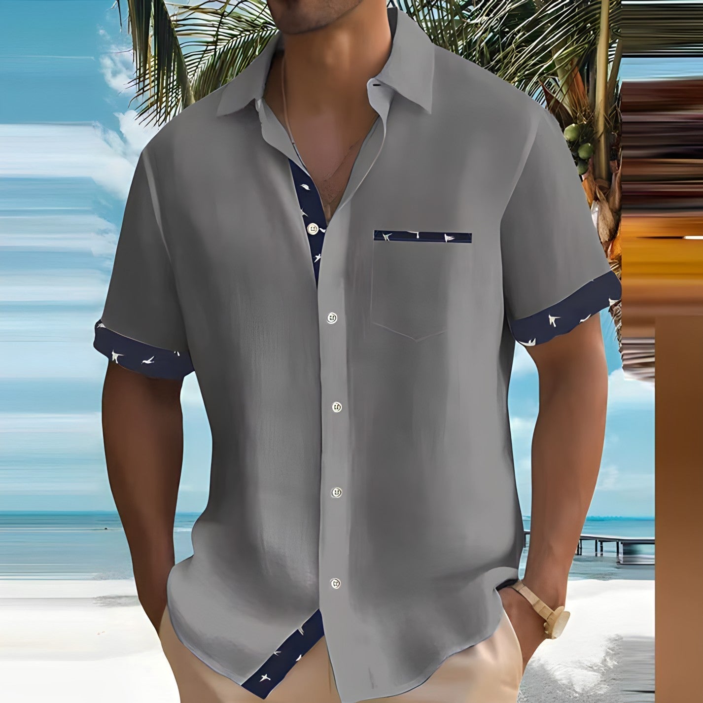Graues Herrenhemd mit blauem Akzent, getragen am Strand. Sommermode, tropisches Design, Kurzarm, lässiger Stil, ideal für Urlaub am Meer.