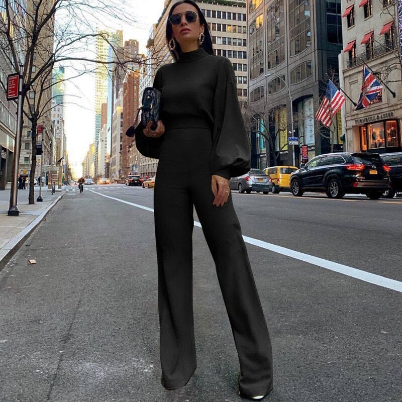 Frau in schwarzem Jumpsuit auf Stadtstraße, hält Clutch, trägt Sonnenbrille, umgeben von Hochhäusern und Autos, USA-Flagge im Hintergrund.