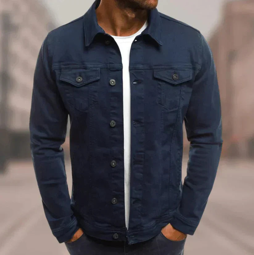Mann in dunkelblauer Jeansjacke und weißem T-Shirt, Hände in den Taschen, urbaner Hintergrund. Modische Herrenbekleidung, lässiger Stil.