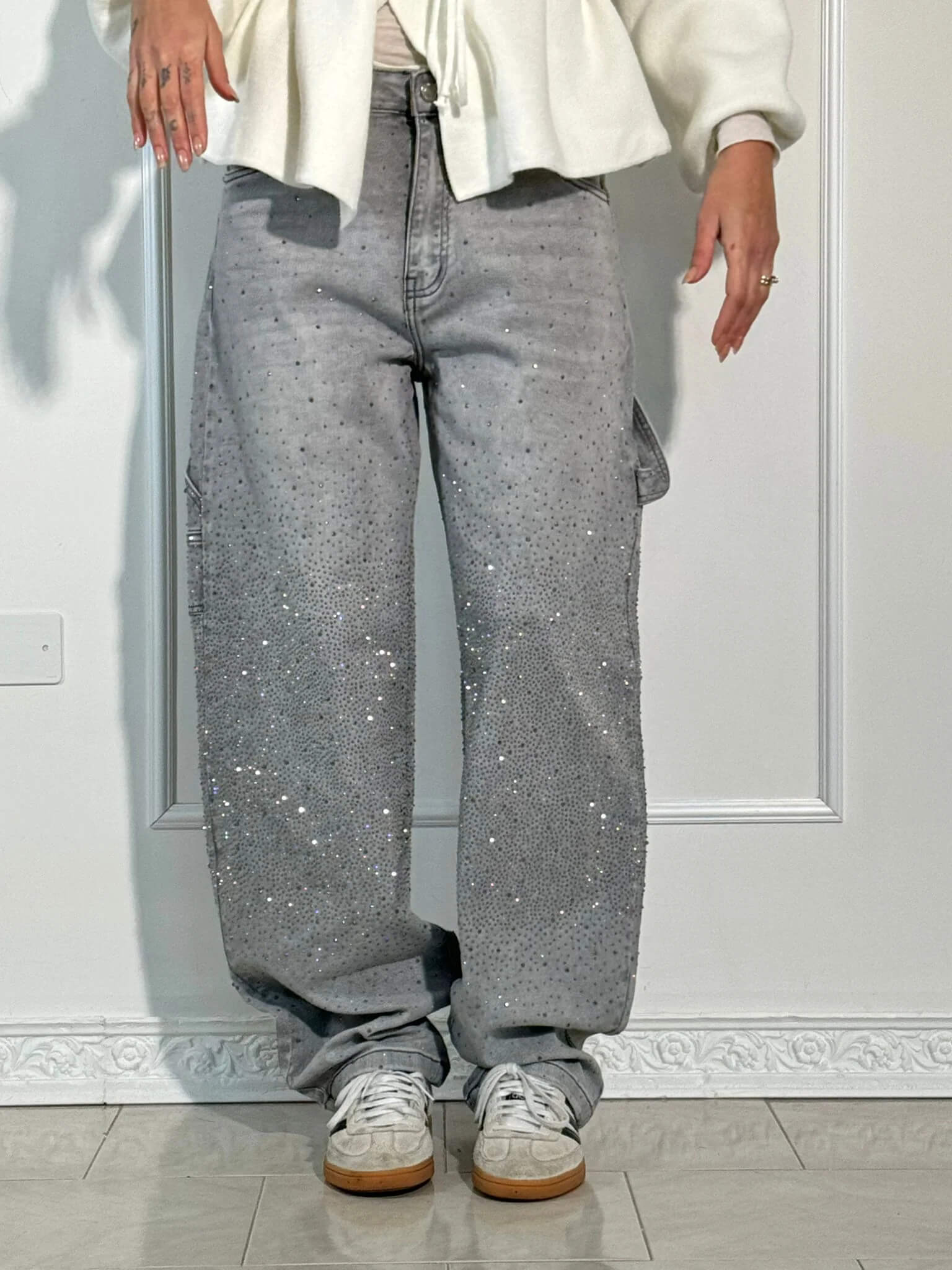 Graue Baggy-Jeans mit funkelnden Strasssteinen, getragen von einer Person mit weißen Sneakers. Modetrend, lässiger Stil, Streetwear.