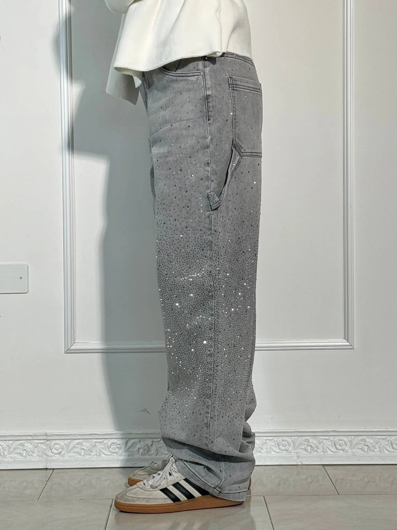 Graue Jeans mit Strasssteinen, seitlicher Tasche und weißen Adidas-Sneakern vor weißer Wand. Modetrend, Streetwear, Herrenmode.