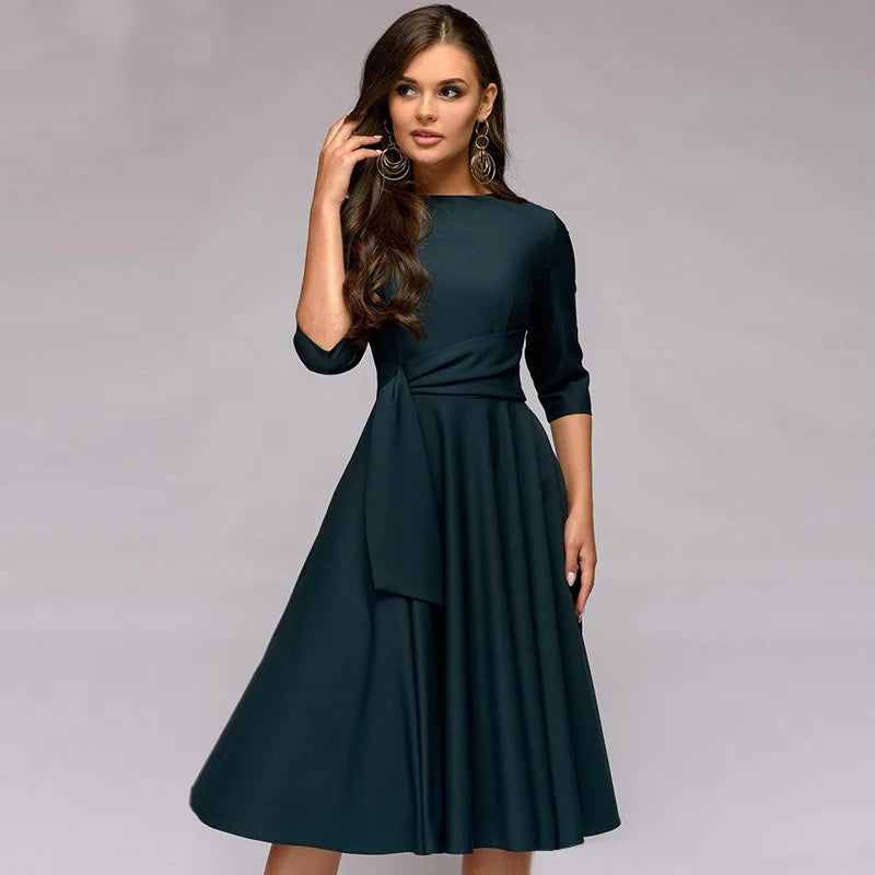 Frau in elegantem, dunkelgrünem Kleid mit Dreiviertelärmeln und Taillengürtel, posiert vor grauem Hintergrund. Mode, Damenbekleidung, Abendkleid.