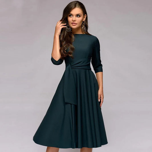 Frau in elegantem, dunkelgrünem Kleid mit Dreiviertelärmeln und Taillengürtel, posiert vor grauem Hintergrund. Mode, Damenkleidung, Abendkleid.